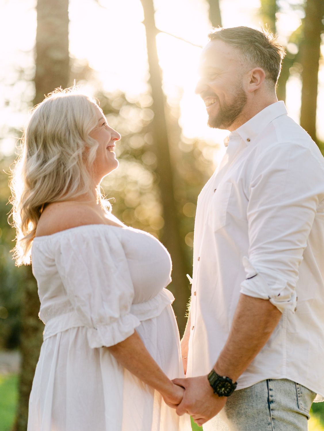 A couple in white outfits holds hands and smiles at each other during a golden hour outdoor photo session.