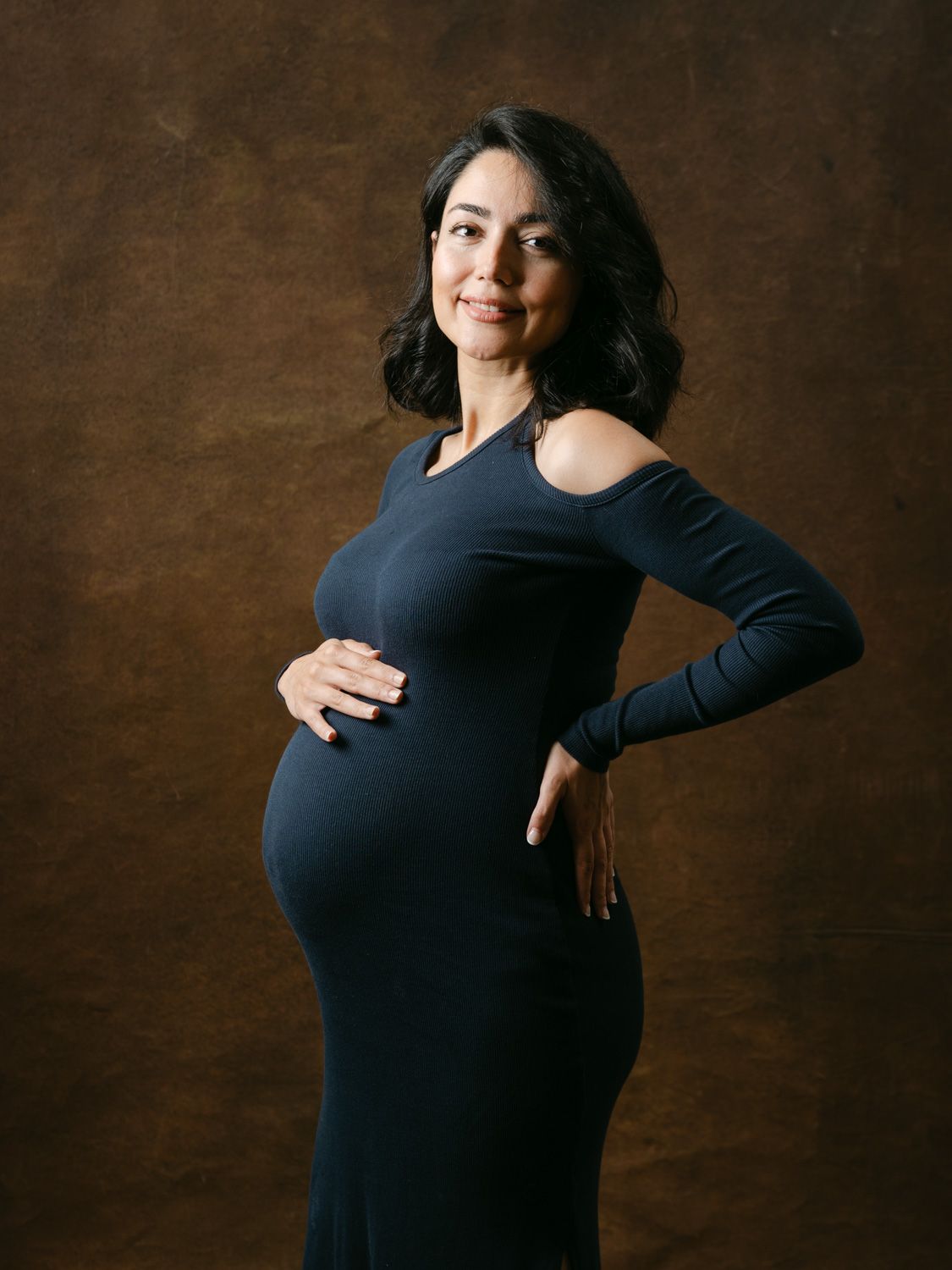 Pregnant woman in navy cold-shoulder dress cradling baby bump, posing elegantly against brown studio backdrop.