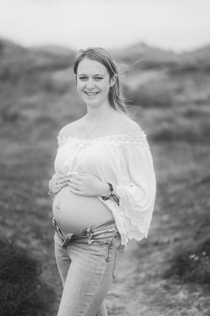 Black and white photo of a smiling pregnant woman in an off-shoulder top and jeans, cradling her baby bump outdoors.