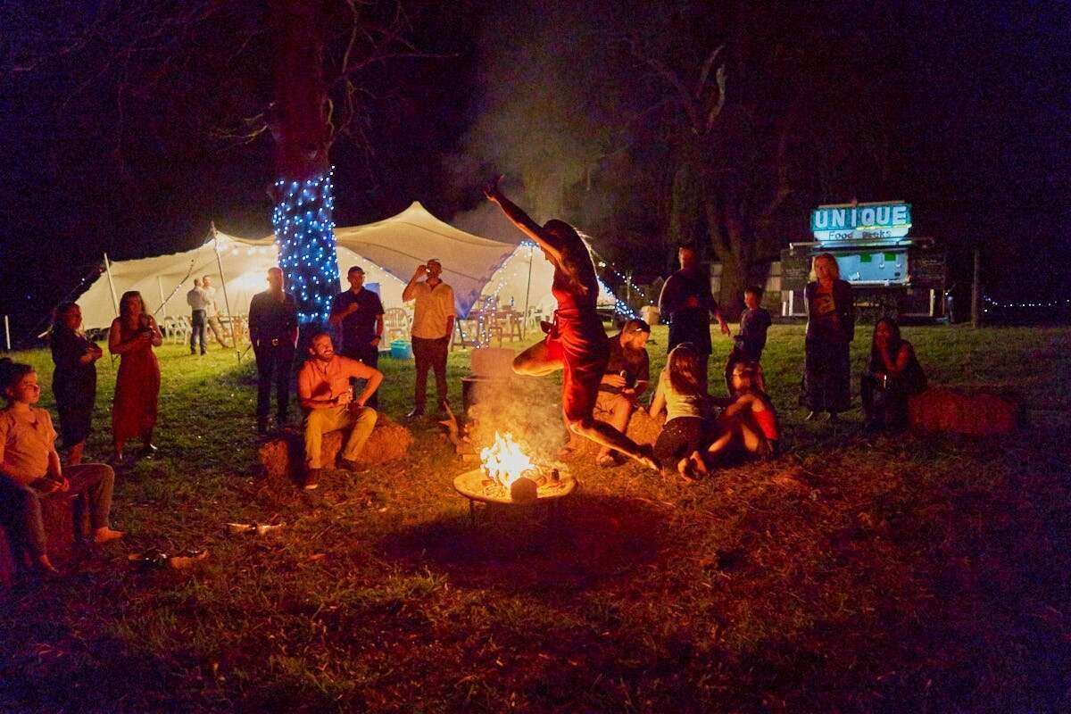 Guests gathered around campfire at night outside illuminated wedding tent with festive lights.