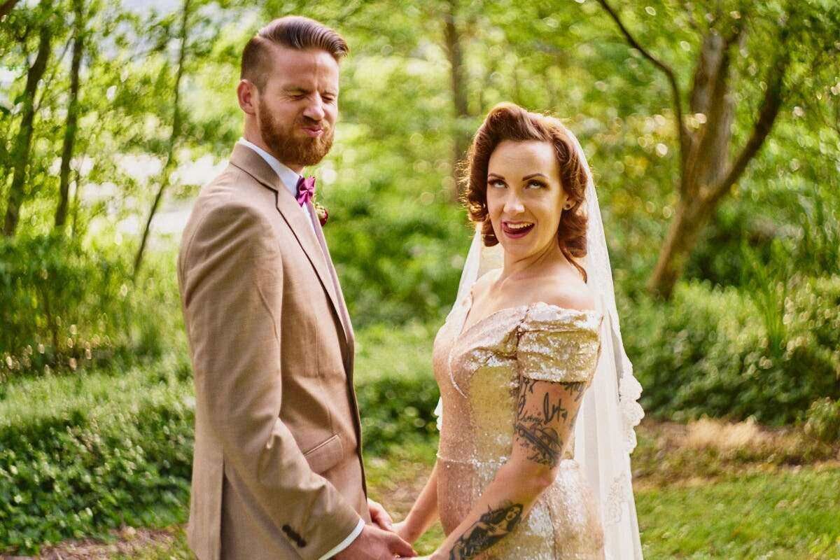 Smiling bride in lace gown and veil holds hands with bearded groom in tan suit at an outdoor wedding.