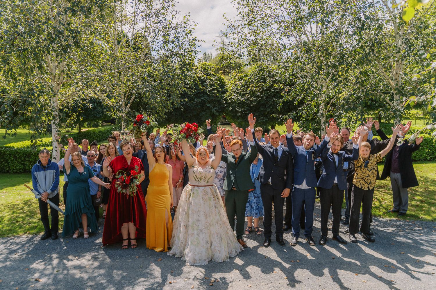 Wedding party celebrating outdoors under green trees, bride and groom surrounded by cheering guests in colorful attire.
