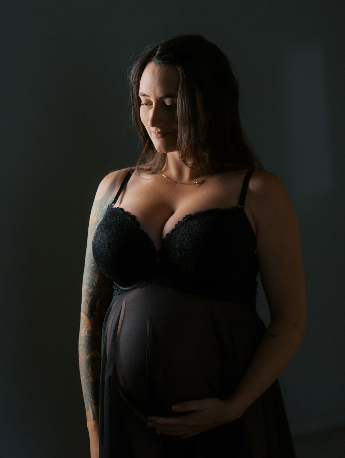 Pregnant woman in black lace lingerie cradling baby bump, tattooed arm, dark moody studio maternity portrait.