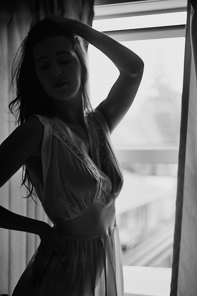 Black and white photo of a woman in lingerie posing by a window with soft backlight.