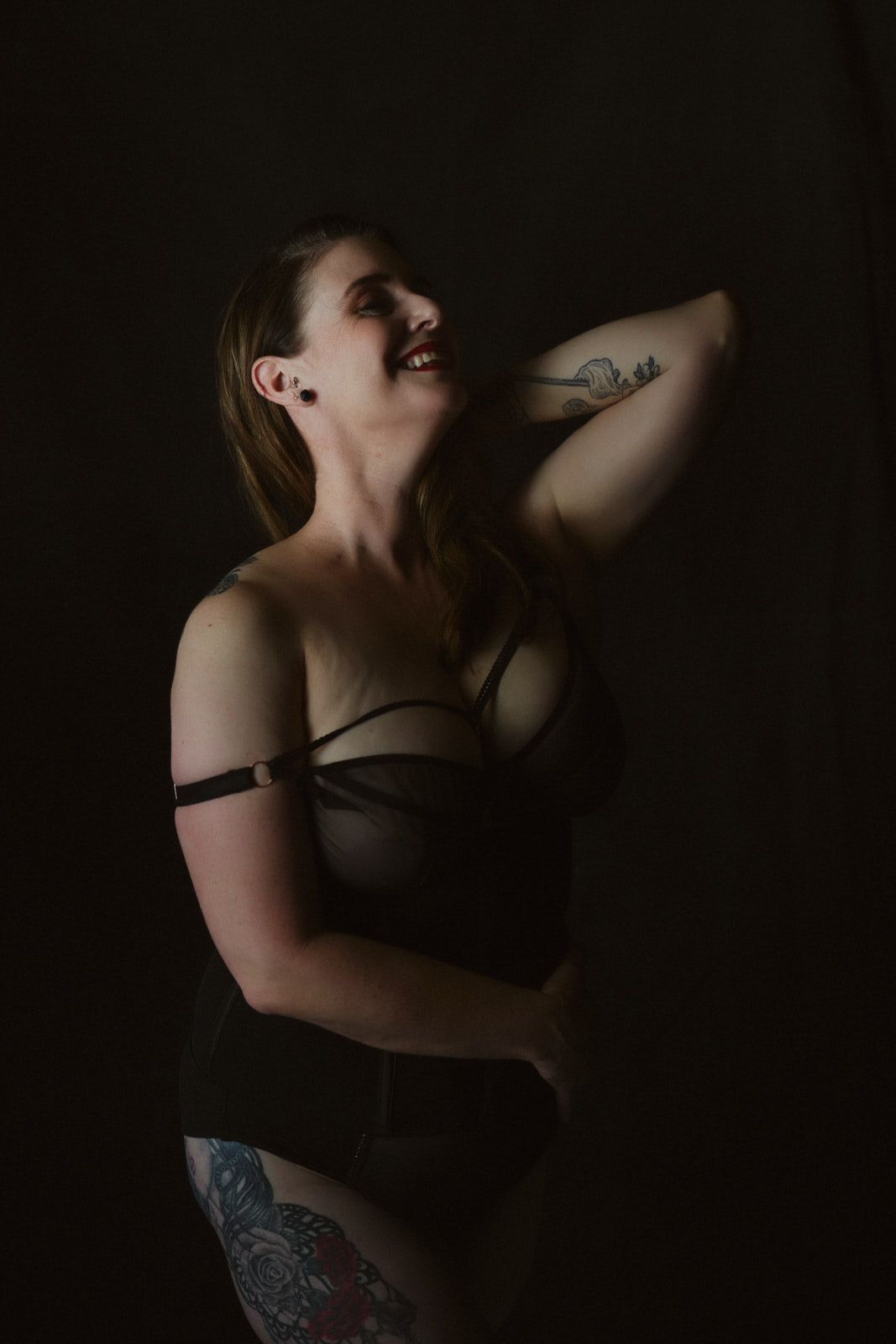 Woman in dark lingerie posing confidently with tattoos visible, shot in dramatic low-key studio lighting.