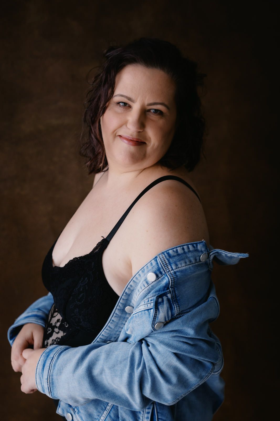 A woman in a black lace bodysuit and denim jacket poses confidently against a dark studio background.