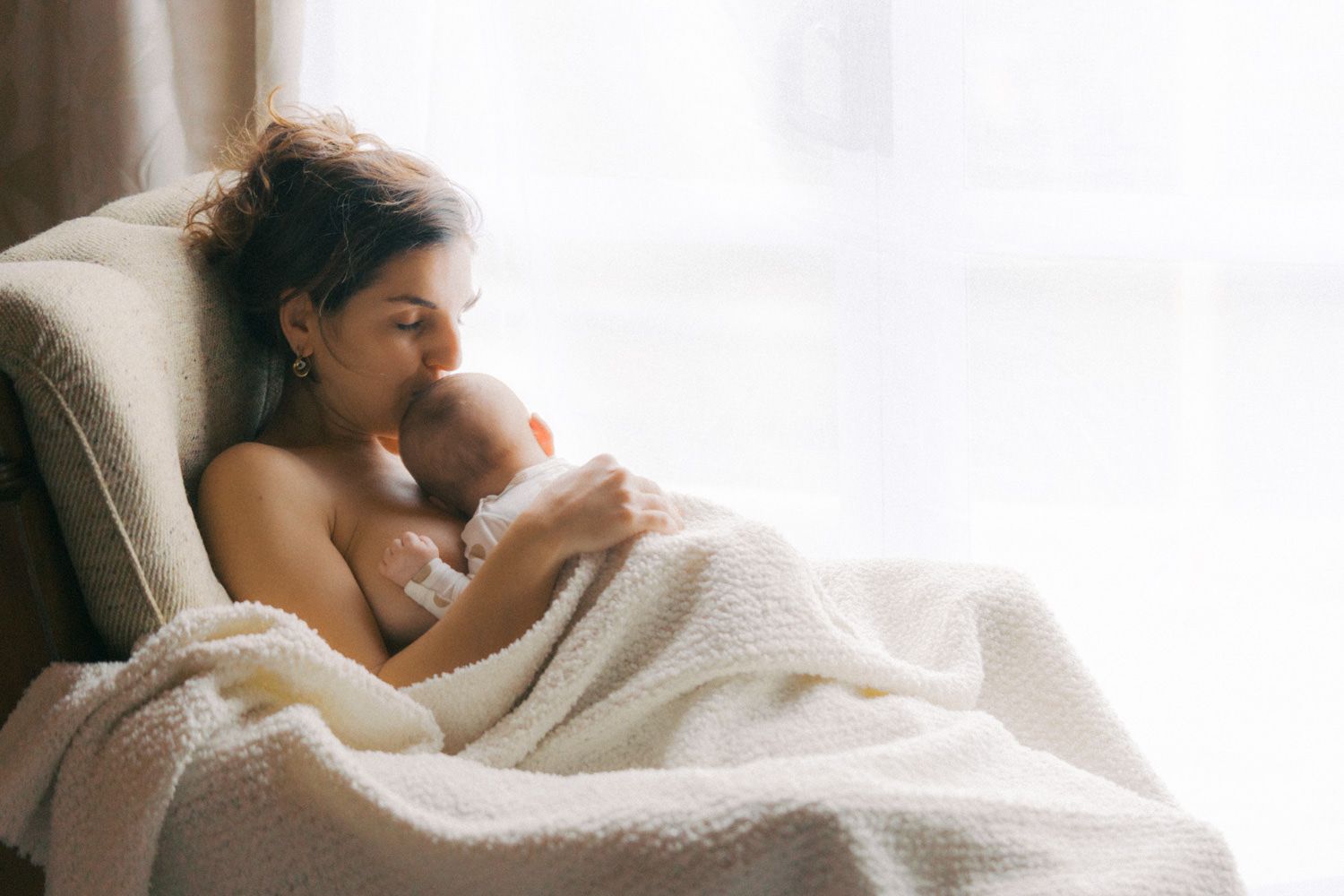 Mother tenderly breastfeeding newborn baby wrapped in soft white blanket while relaxing in armchair by window.