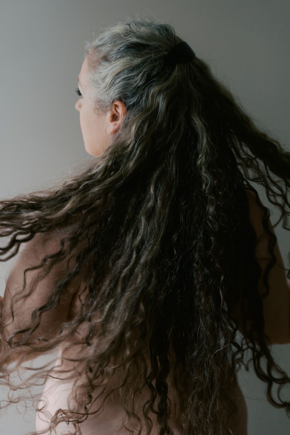 Woman with long, flowing gray and dark curly hair viewed from behind against a neutral background.