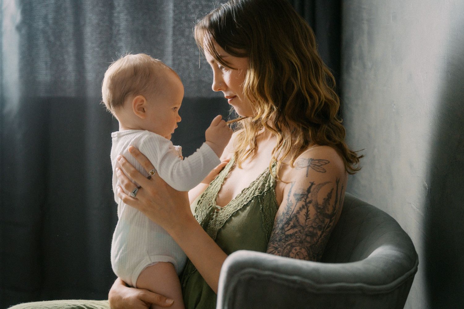 A tattooed mother smiles lovingly at her baby while seated in a gray chair near curtains.