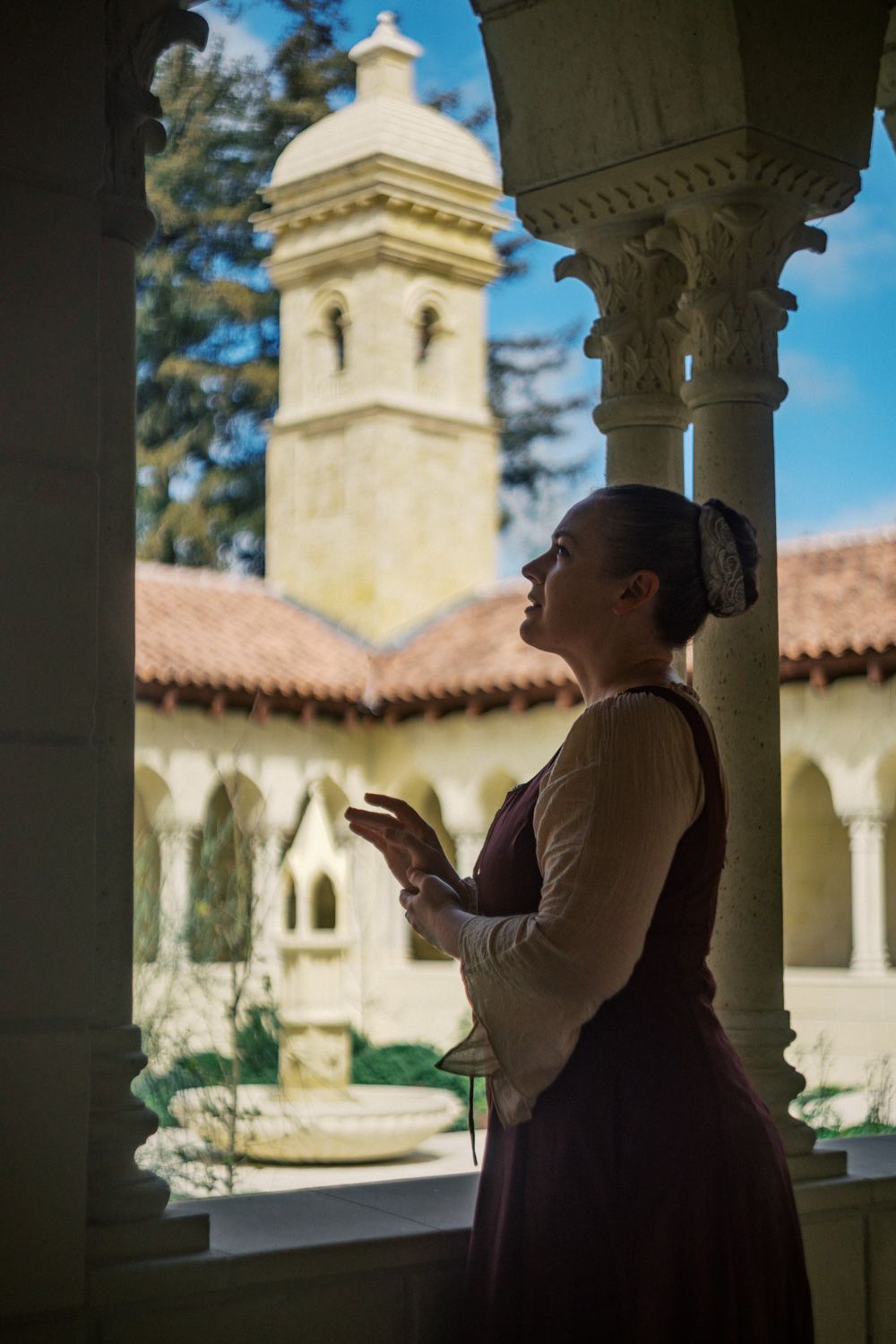 Woman in period costume gazes upward in a sunlit Mediterranean-style courtyard with ornate columns and a bell tower.
