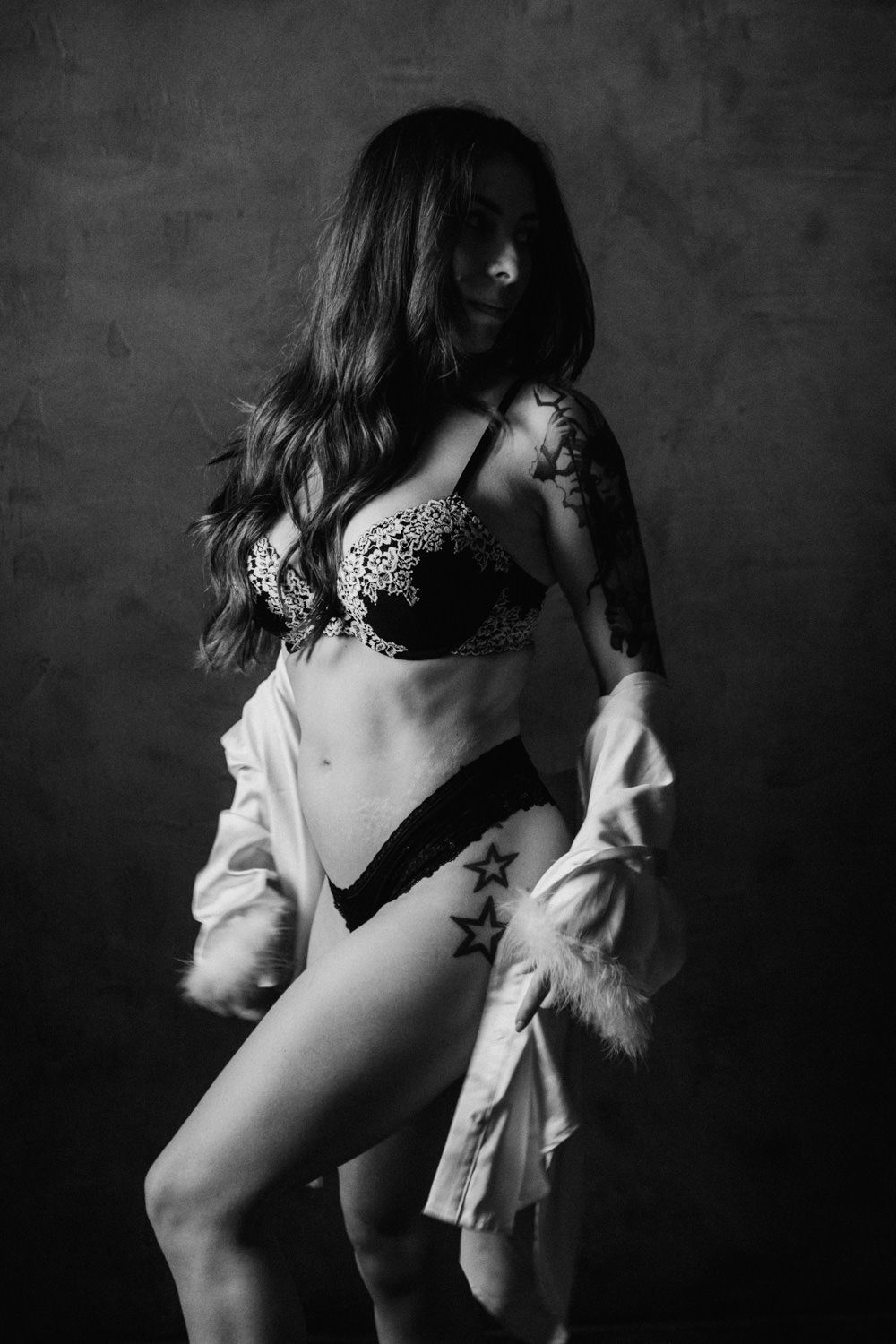 Black and white boudoir photo of a woman in lace lingerie and open white shirt with star tattoos on her hip.