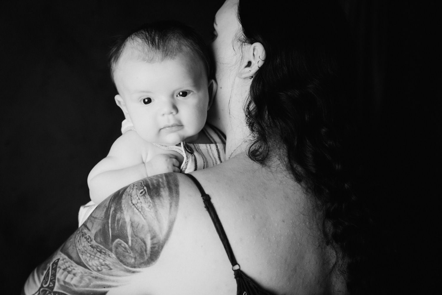 Black and white photo of a tattooed mother holding her newborn baby against a dark background.