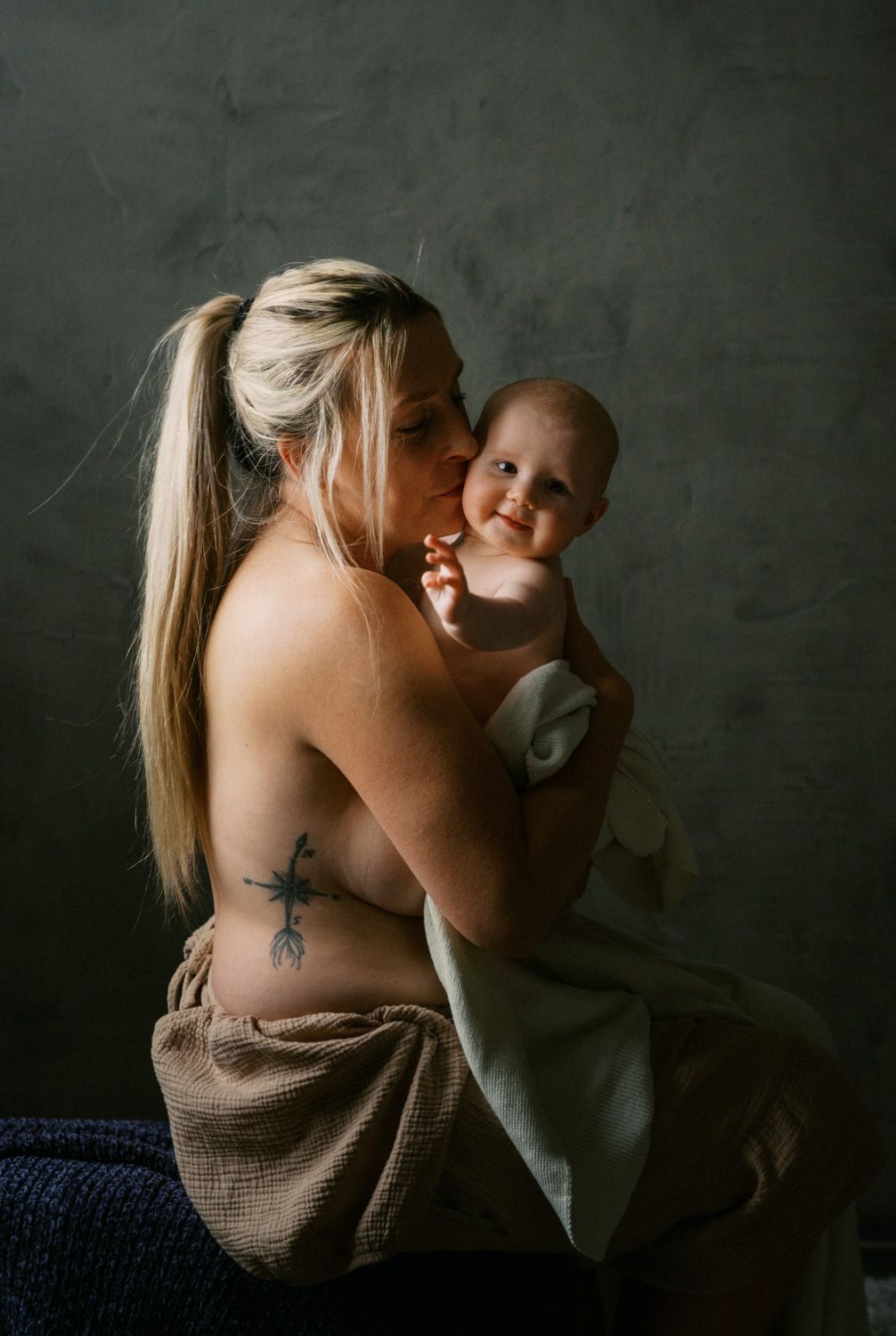 A mother with a blonde ponytail and tattoo kisses her smiling baby wrapped in a soft cloth against a gray backdrop.