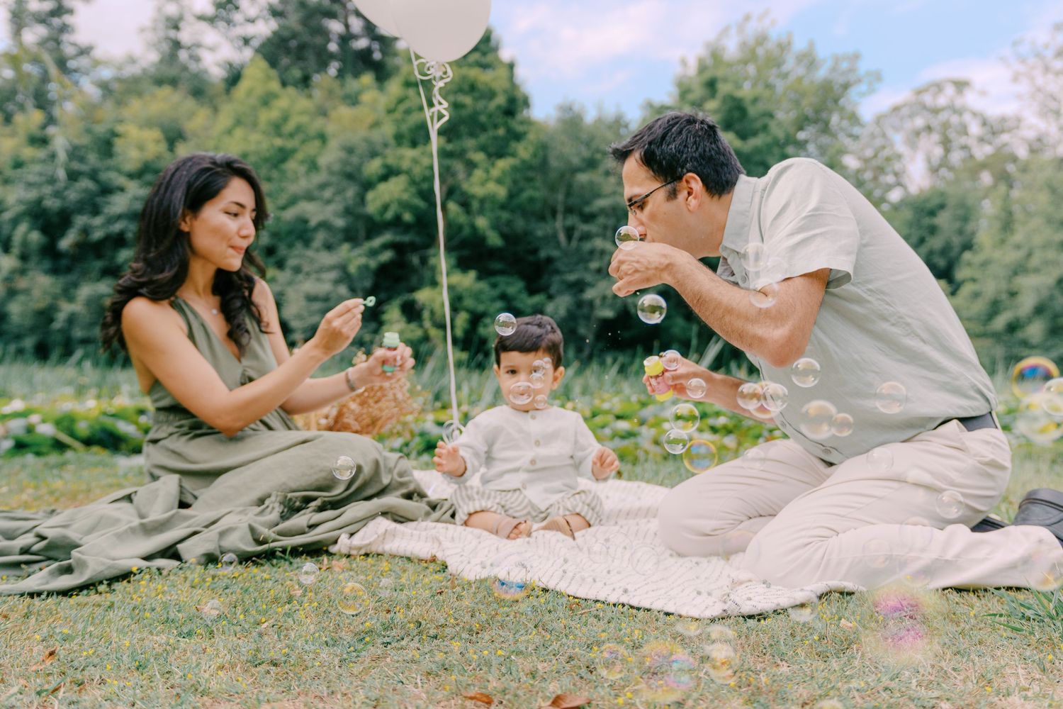 Family enjoying a outdoor picnic on a blanket in a lush green park, parents blowing bubbles for their baby.