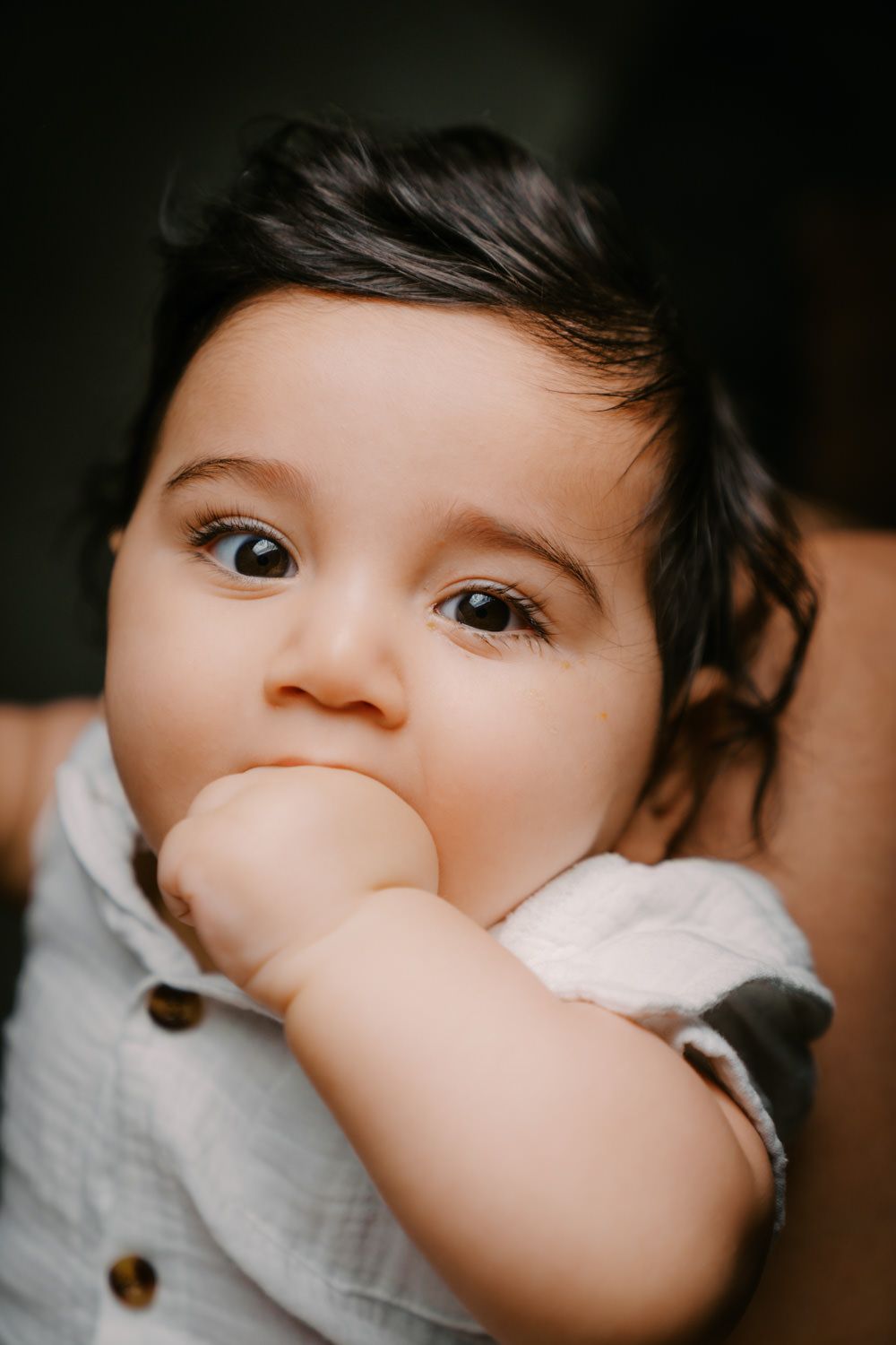 Adorable baby with dark hair and big brown eyes chewing on their fist, wearing a light gray button-up shirt.