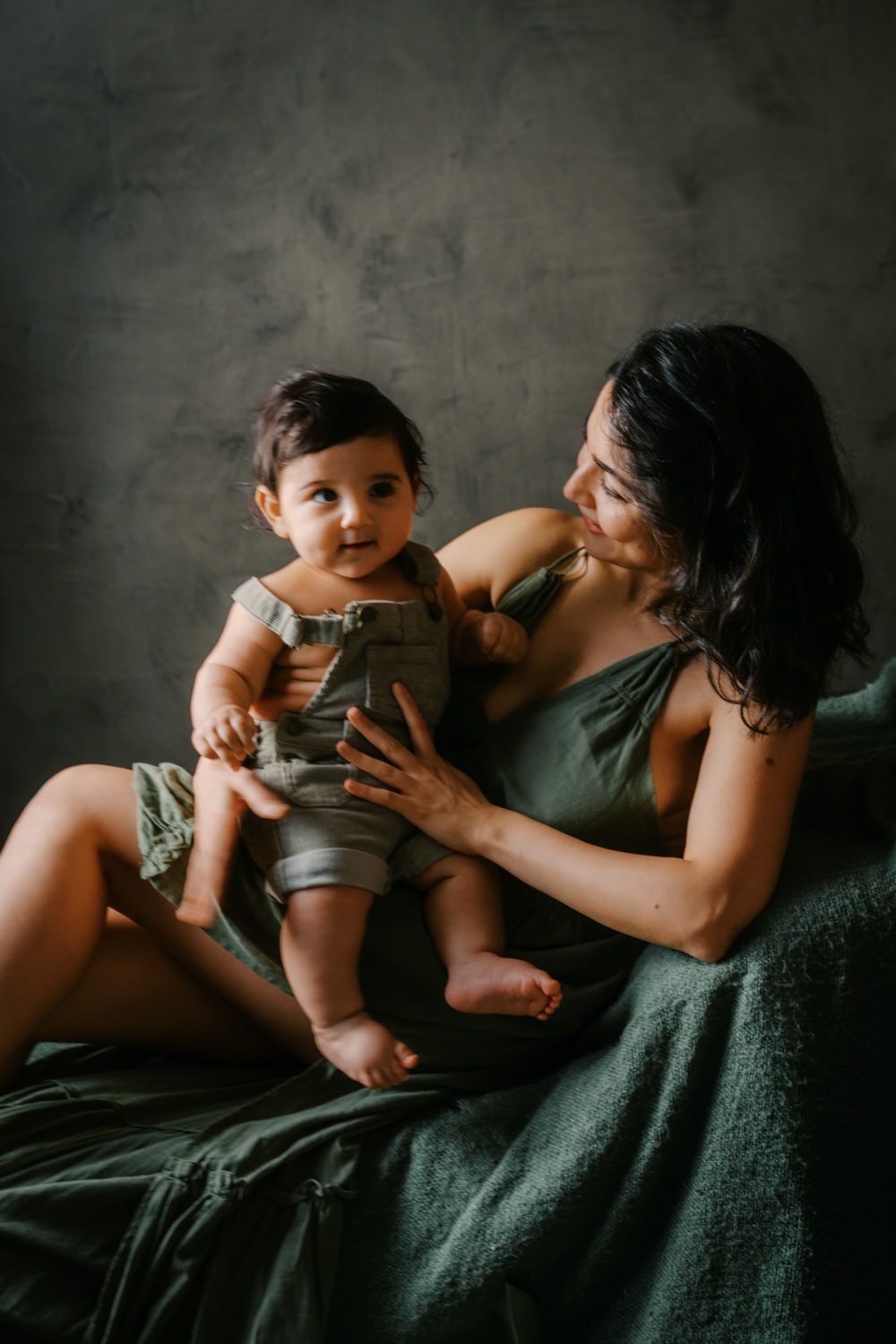 A mother in a green dress holds her baby in olive overalls while sitting on a draped chair against a gray backdrop.