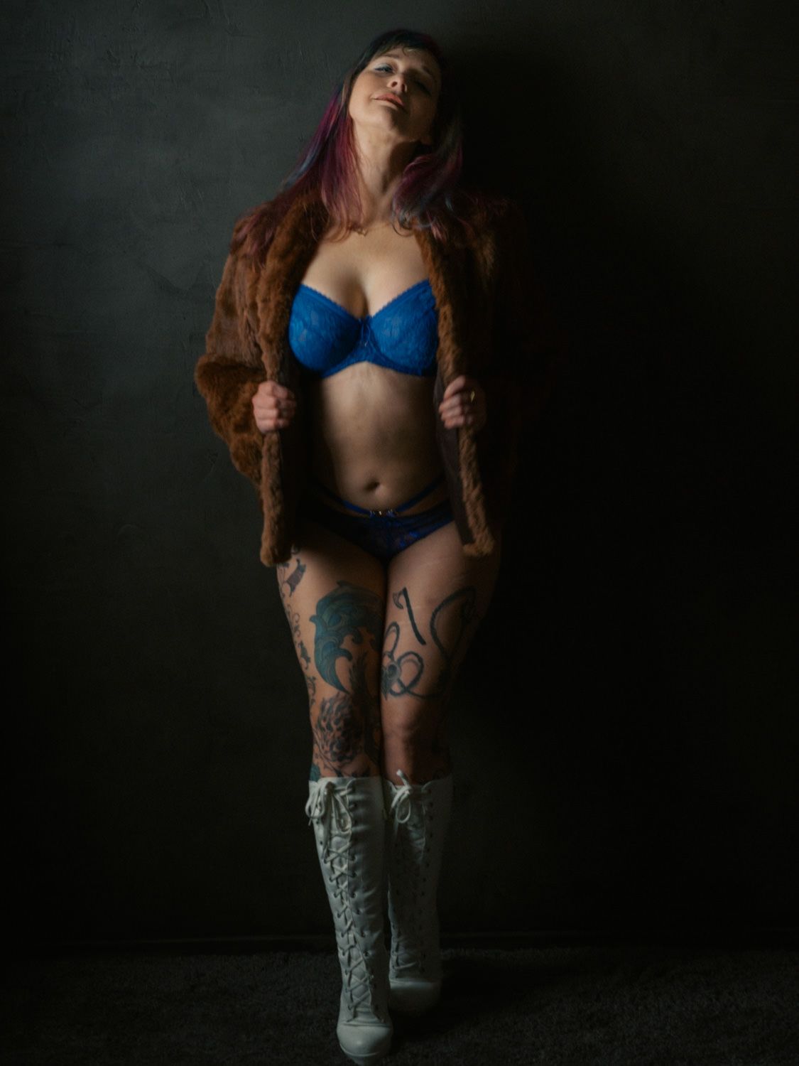 Woman in blue lingerie and fur coat with tattoos and white boots poses confidently against dark background.