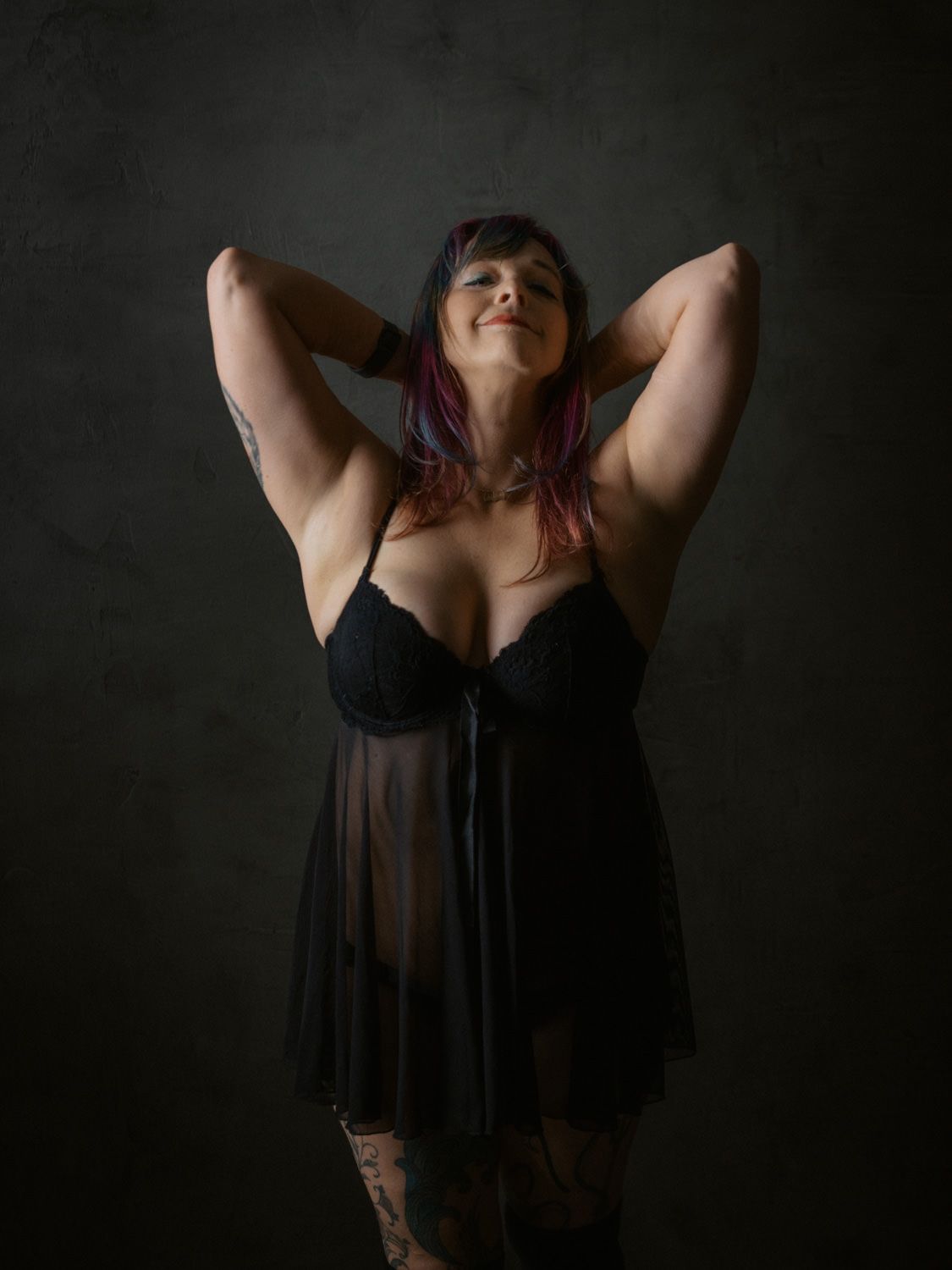 Woman in black sheer lingerie poses with hands behind head against dark studio backdrop, showing tattoos and colorful hair.
