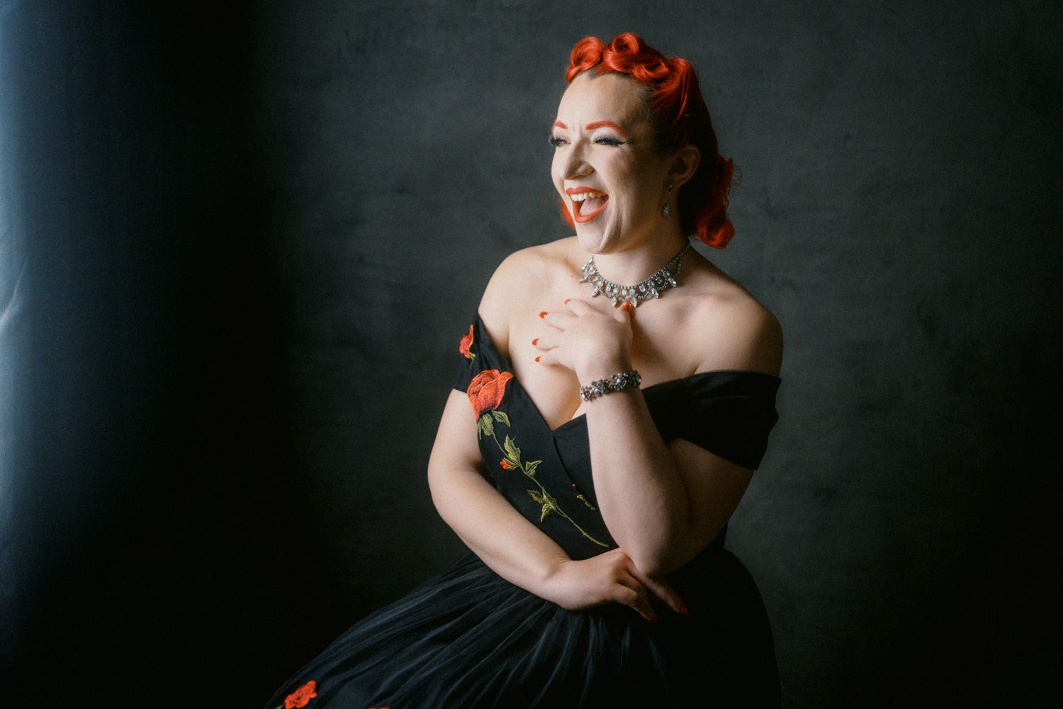 Woman in black off-shoulder dress with red floral embroidery, red pin-up hair, smiling playfully against dark background.