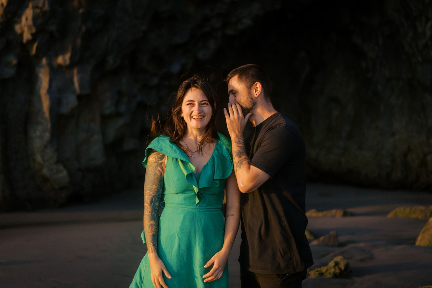 Pregnant woman in teal dress smiles as man whispers in her ear near rocky cave backdrop.