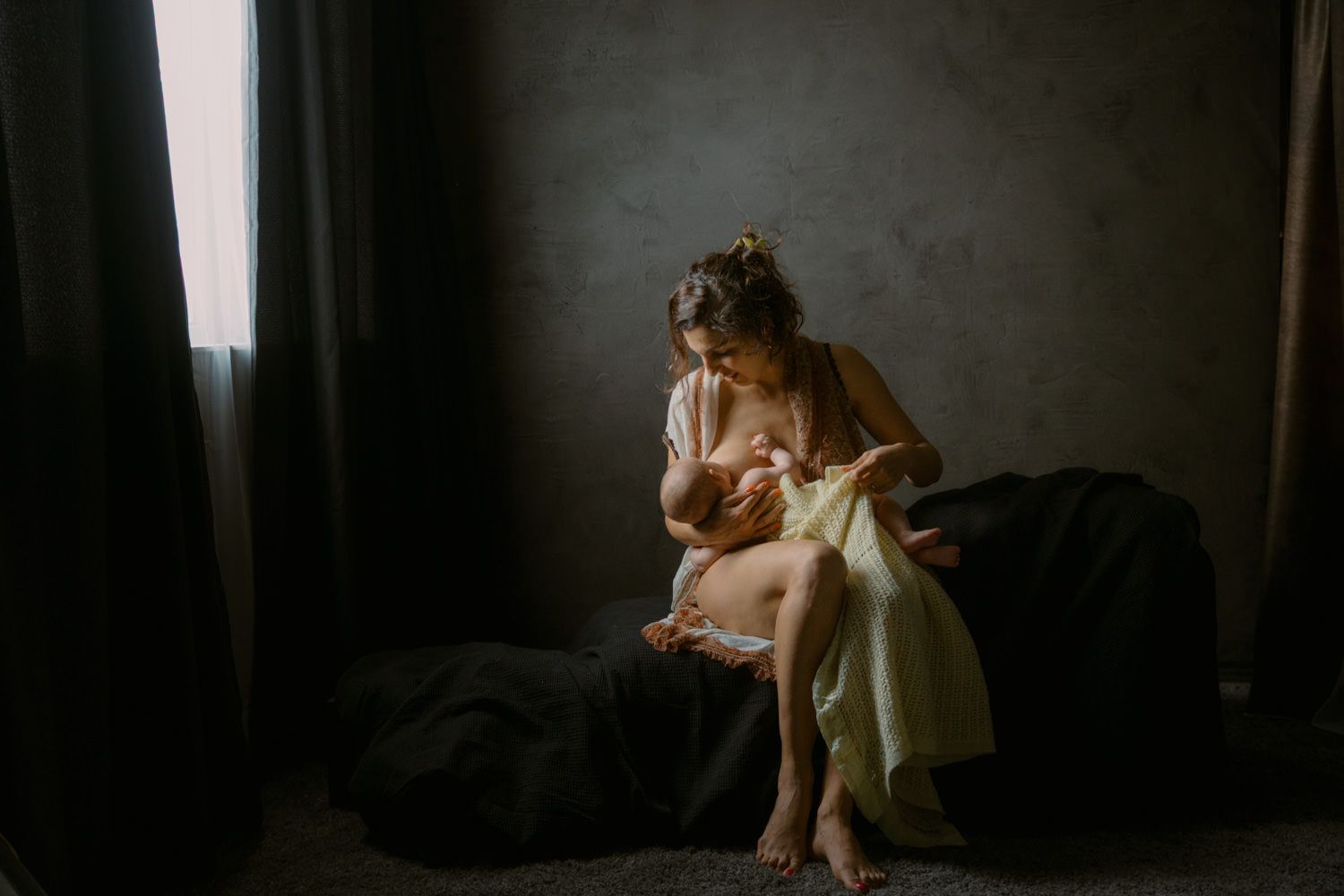 A mother breastfeeding her infant in soft natural light in a dimly lit room.