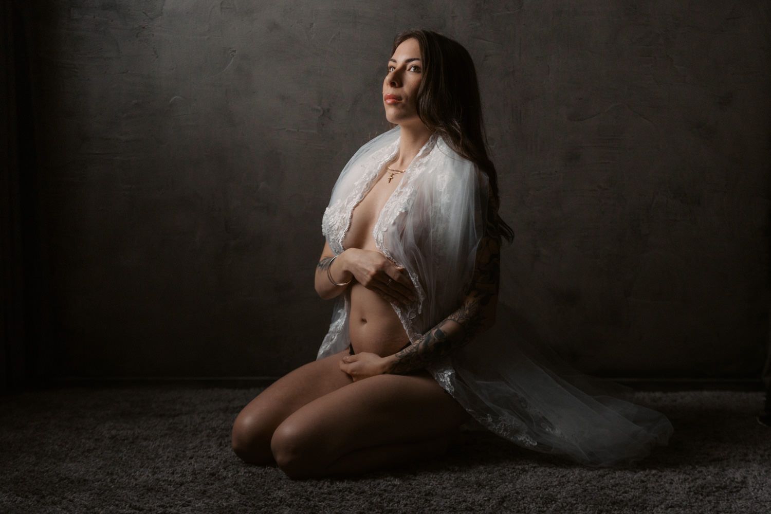 A woman in an open white robe poses gracefully on her knees against a dark textured wall in a boudoir photo.