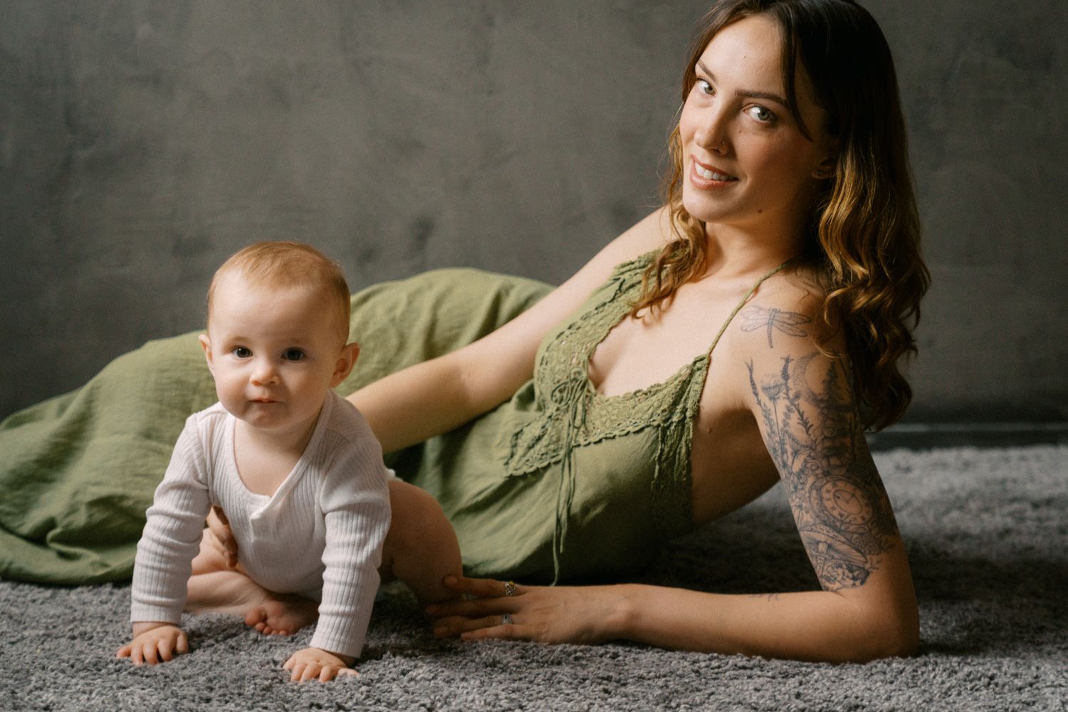 Smiling tattooed mother in green dress lying on carpet beside crawling baby in white onesie against gray wall.