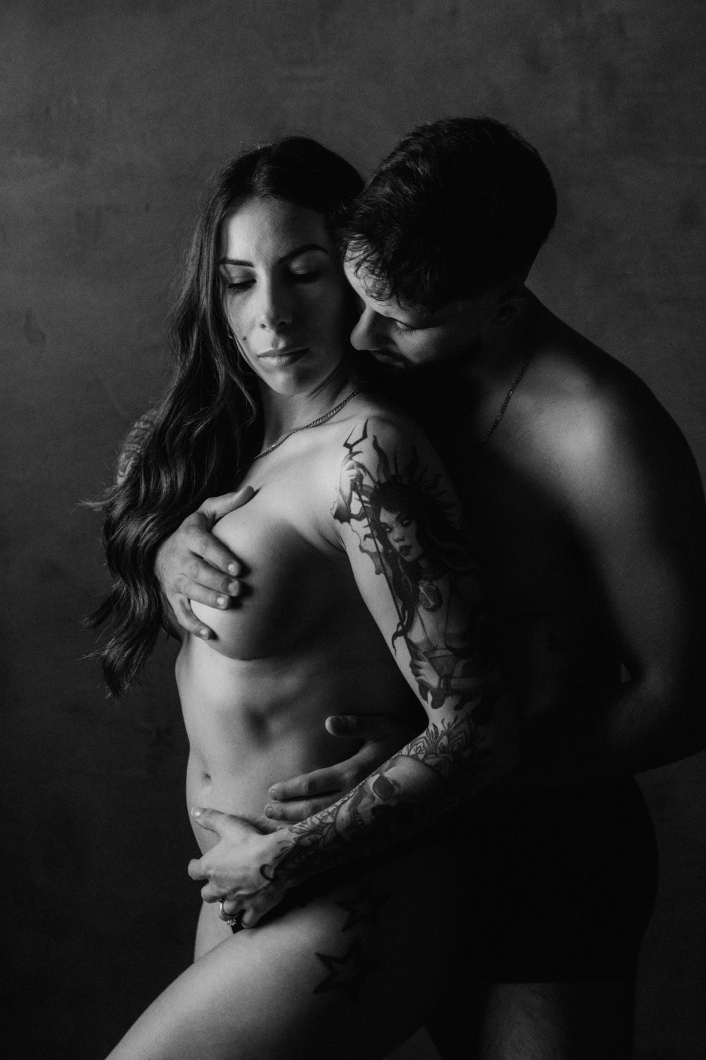 Black and white maternity photo of a couple embracing, woman with tattoos posing nude while partner holds her from behind.