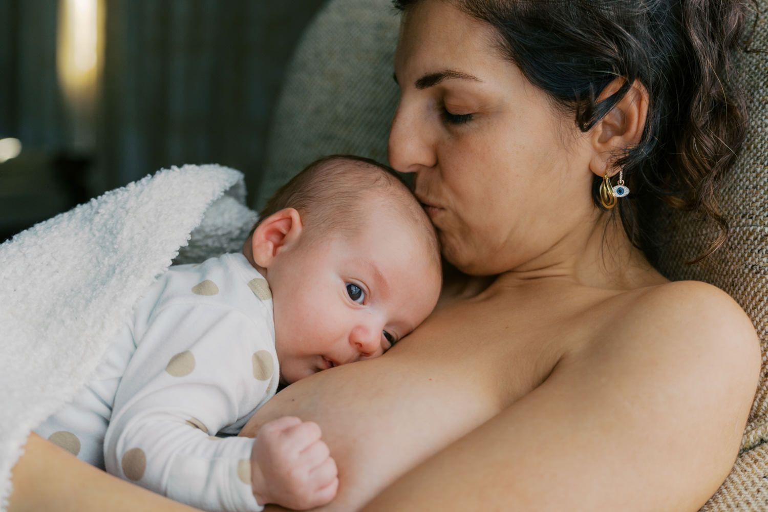 A mother tenderly kisses her newborn baby while cradling them skin-to-skin in a cozy chair.