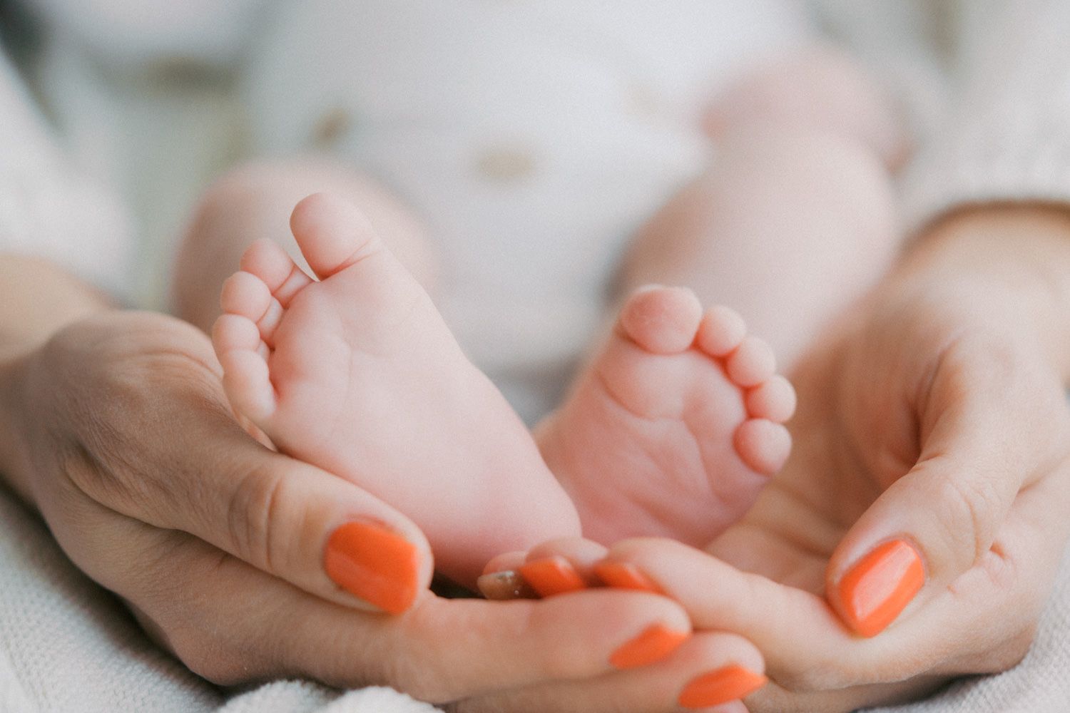 Close-up of a mother's hands with orange nails gently cradling tiny newborn baby feet.