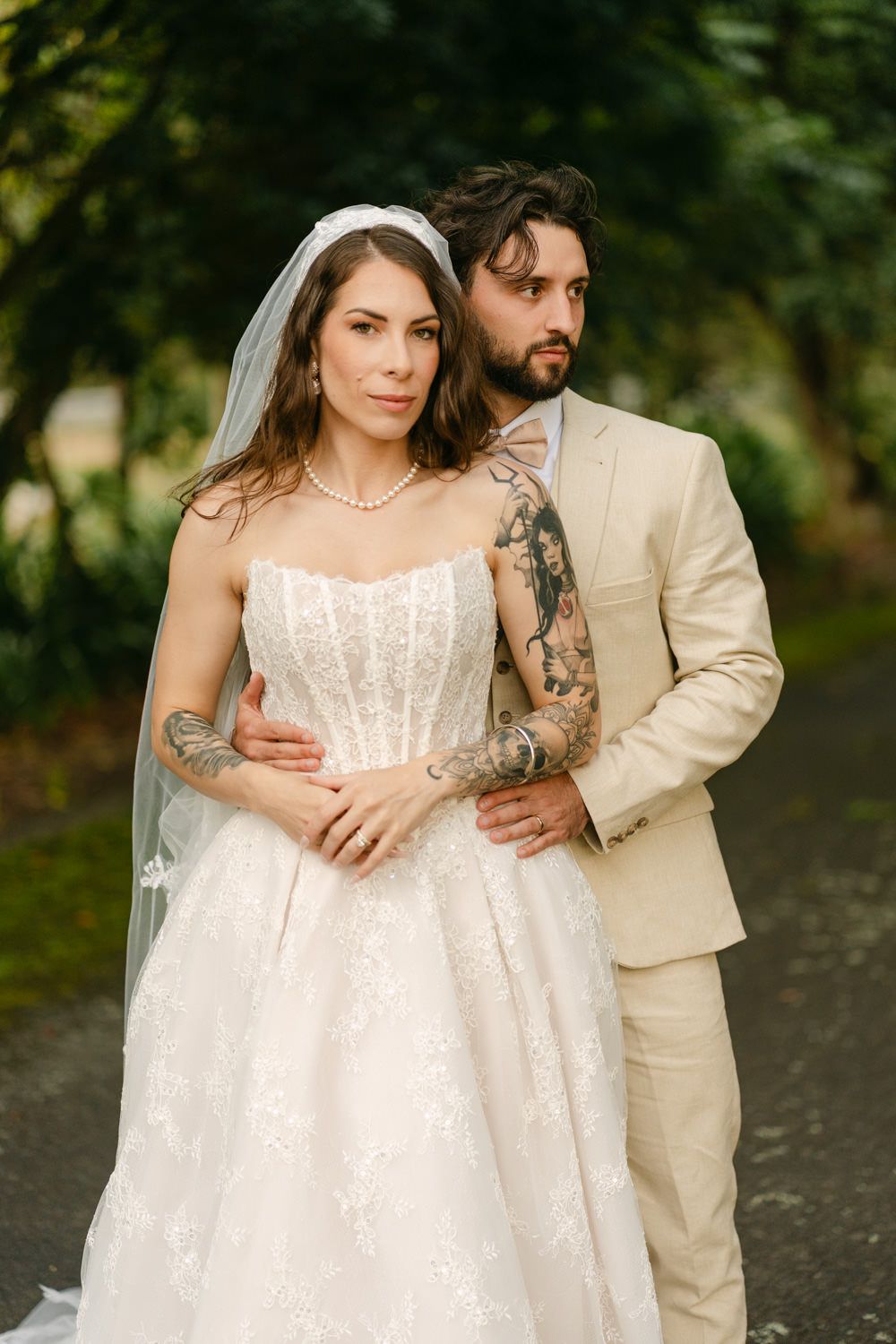 Bride in strapless lace gown with veil and groom in tan suit with tattoos pose outdoors among green trees.