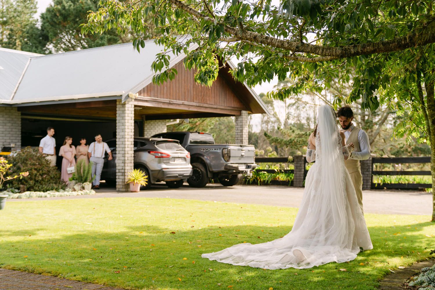 Bride in elegant white gown with long train stands on green lawn outside rustic venue with guests in background.
