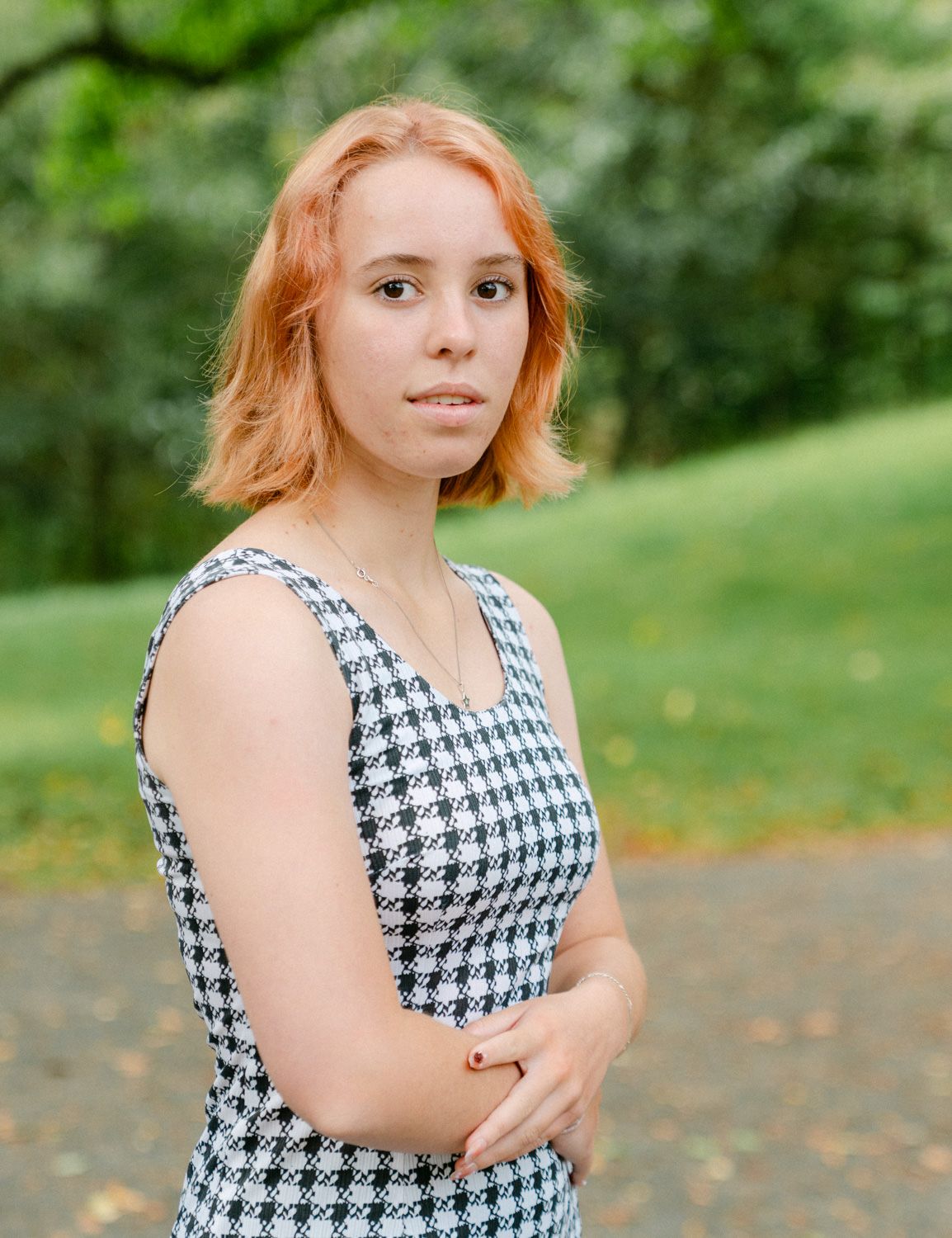 Young woman with red hair wearing a houndstooth dress stands in a green park, arms crossed.