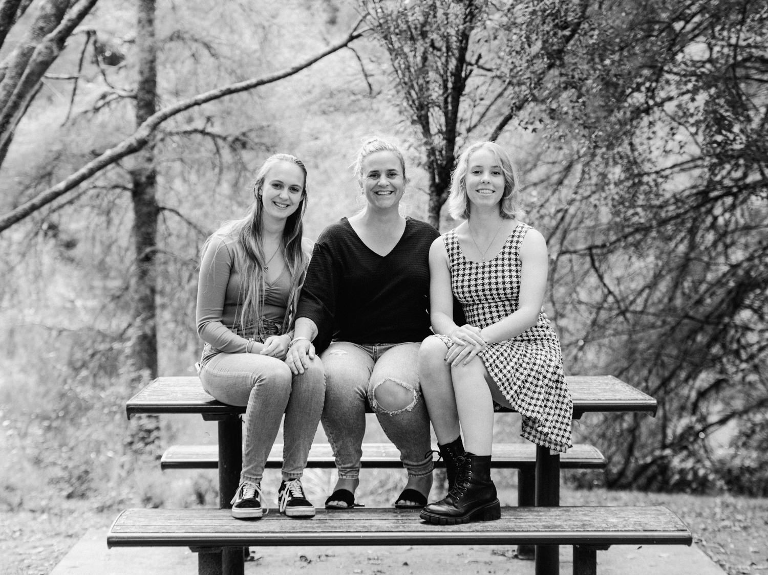 Three young women sitting together on a picnic table bench outdoors, smiling, in a black and white photo.