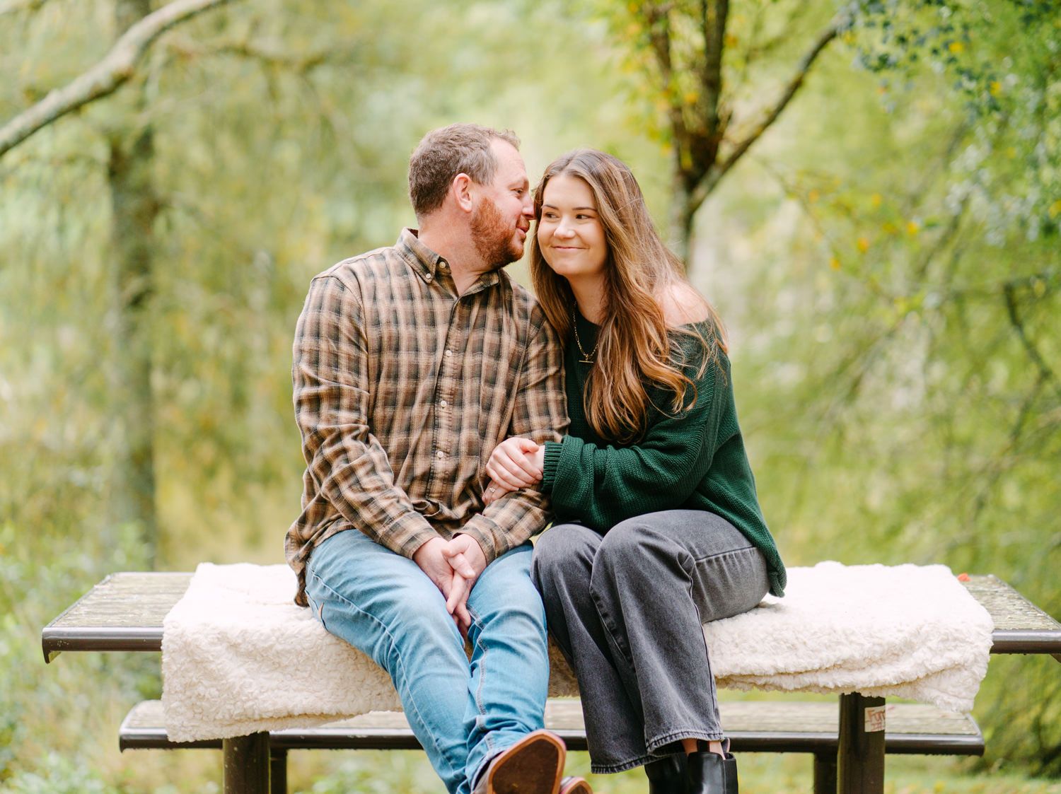 Couple sitting on a wooden bench in a lush green forest, smiling and holding hands in a romantic outdoor setting.