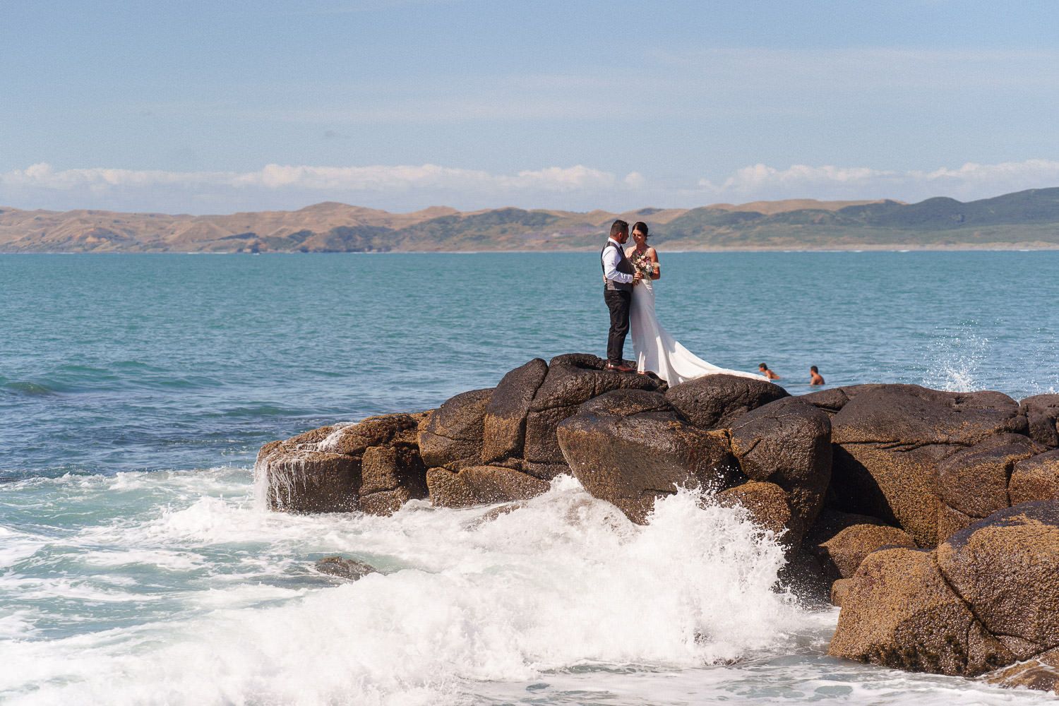 Bride and groom embracing on rocky shoreline with ocean waves crashing around them and mountains in the background.