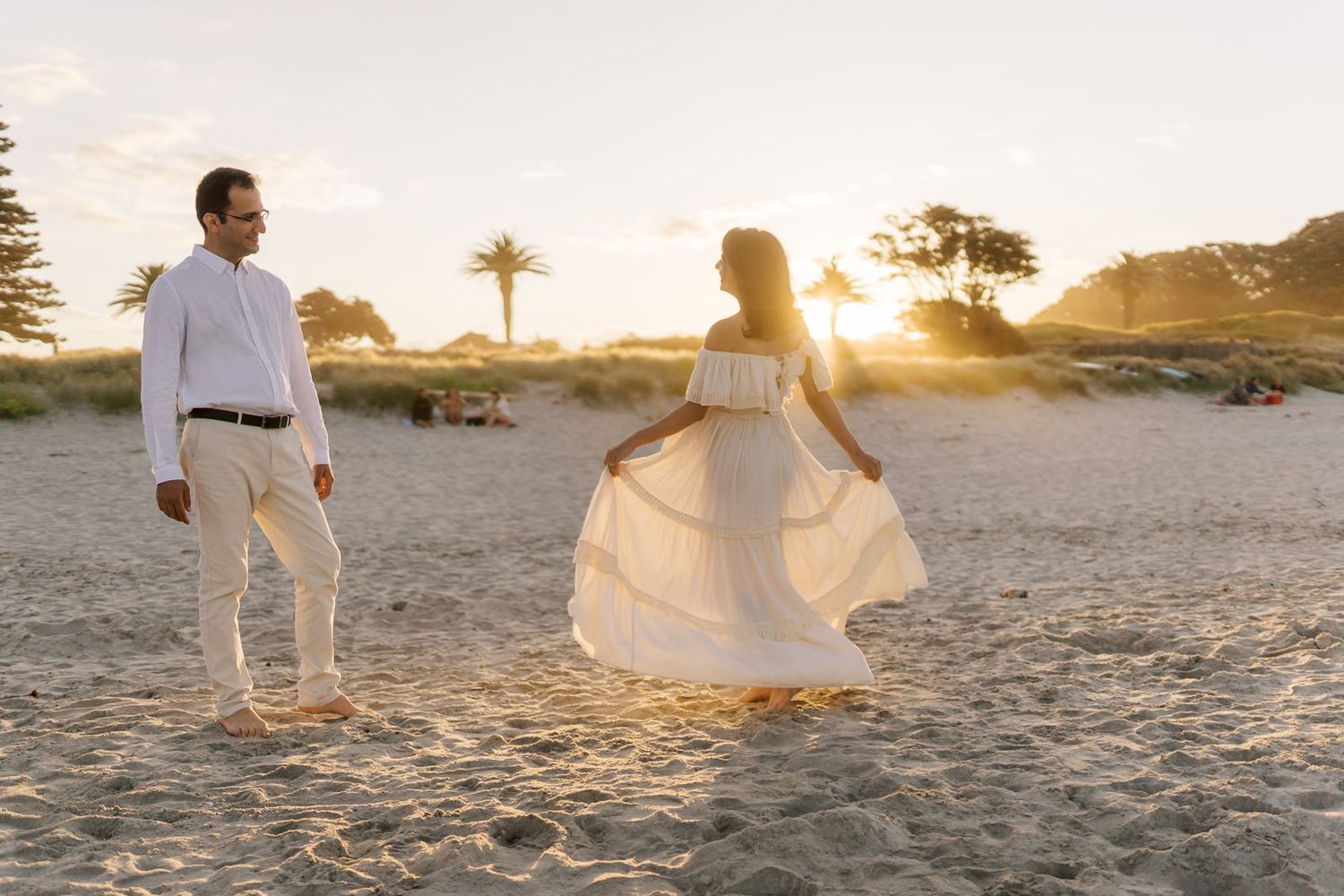 Couple in white attire on sandy beach at golden sunset, woman twirling dress, palm trees in background.