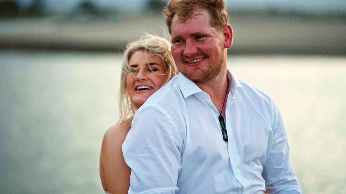 Happy couple in white shirts laugh and embrace near a calm waterfront at golden hour.