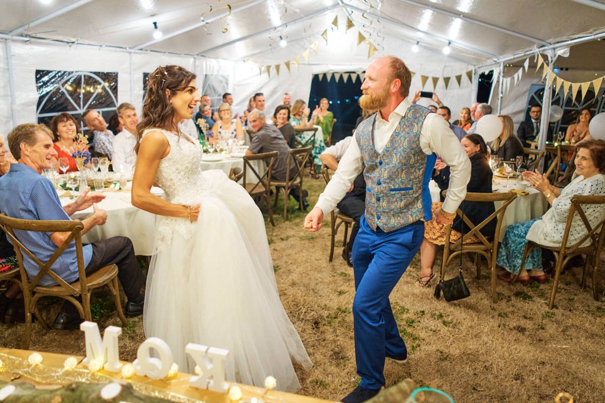 Bride in white gown and groom in blue suit share first dance at elegant outdoor wedding reception under a marquee tent.