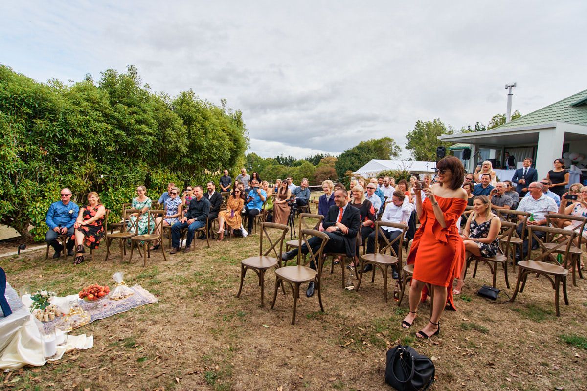 Outdoor wedding ceremony with guests seated on wooden chairs in a garden, officiant in orange dress addressing the crowd.