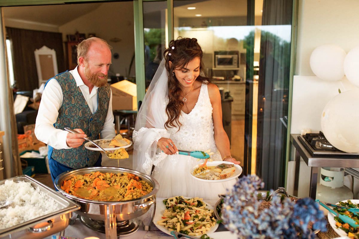 Bride and groom serving themselves at a wedding buffet with colorful dishes spread across a decorated table.