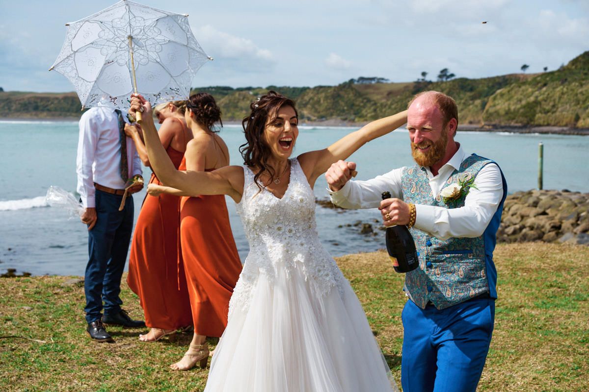 Bride and groom celebrating outdoors near ocean, popping champagne with bridesmaids in orange dresses on sunny wedding day.