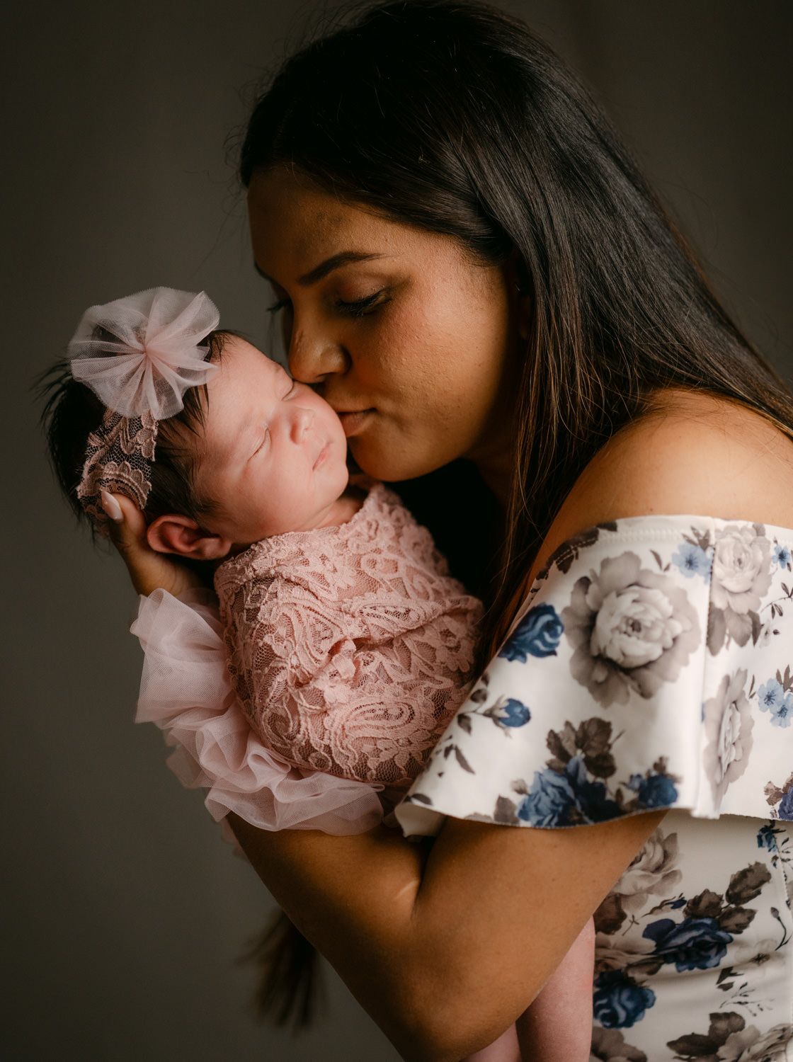 A mother tenderly kisses her newborn baby girl dressed in pink lace wrap with a bow headband.