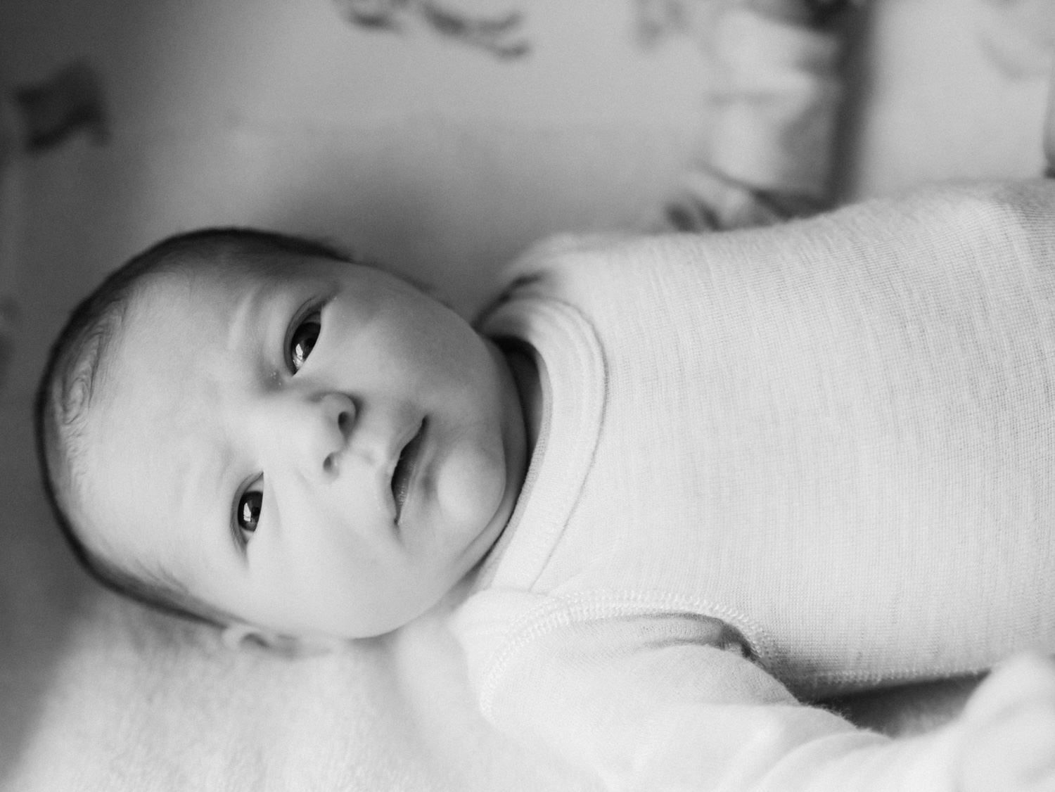 Black and white portrait of a newborn baby wrapped in white swaddle, gazing upward with wide eyes.