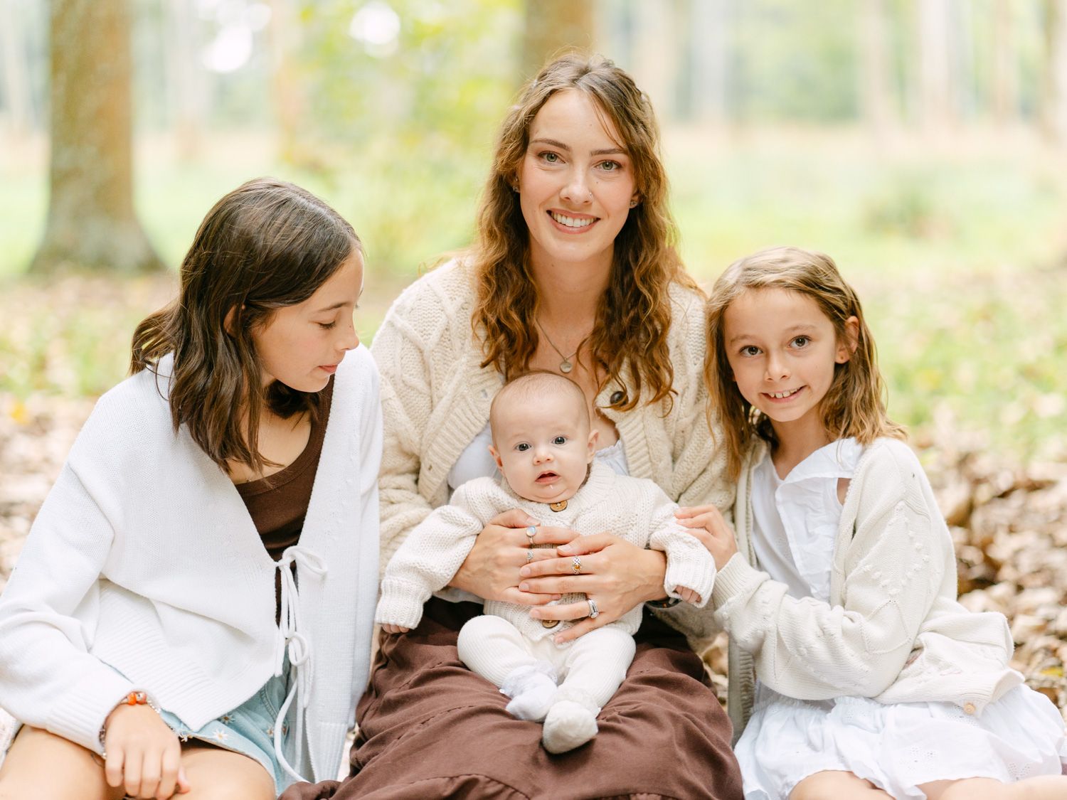 A smiling mother sits outdoors with her baby and two daughters in a warm, wooded setting.