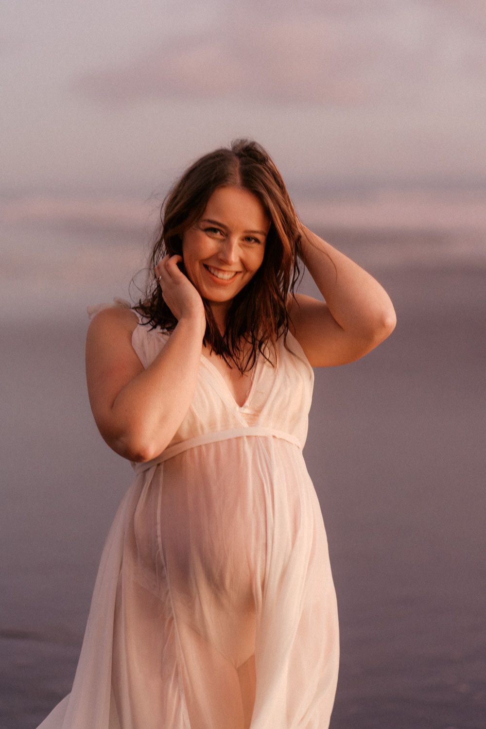 Smiling pregnant woman in flowing white sheer dress poses at beach during golden hour sunset.