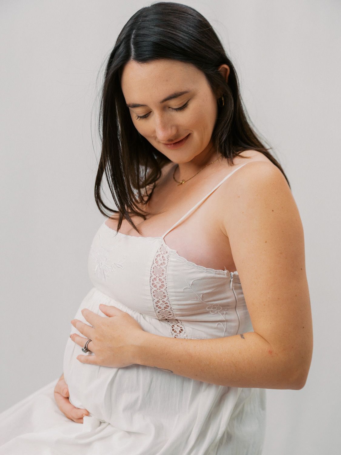 Pregnant woman in white lace dress cradling baby bump, looking down with a gentle smile, maternity photo.