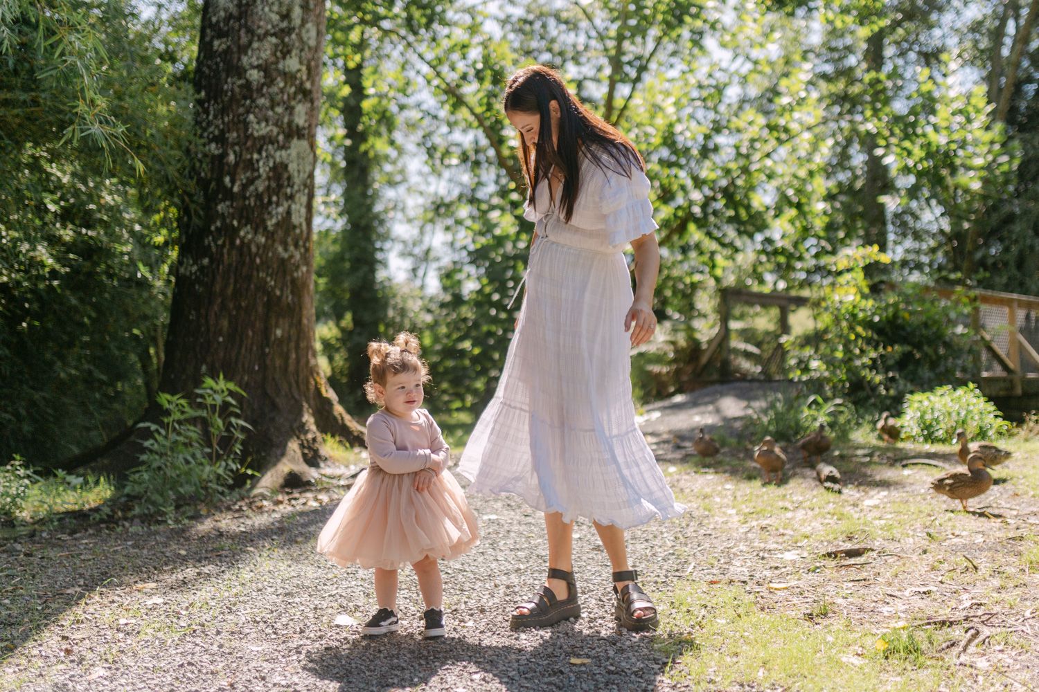 A mother in a white dress holds hands with her toddler daughter in a pink tutu on a sunny garden path.