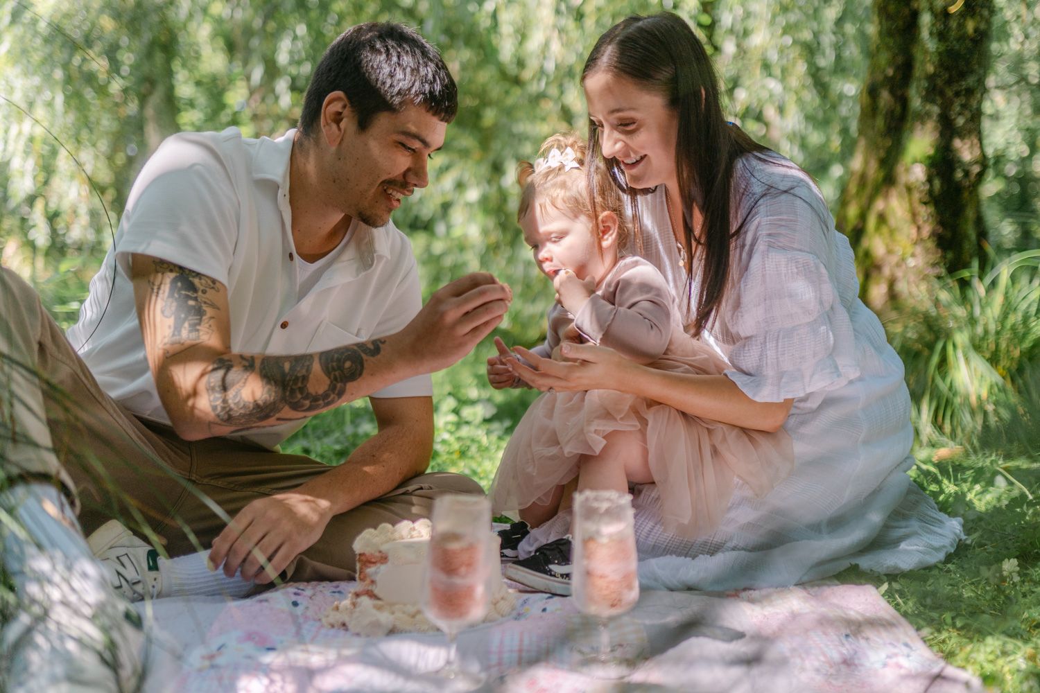 Family enjoying a sunny outdoor picnic on a blanket, with tattooed dad and mom sharing a sweet moment with their toddler.