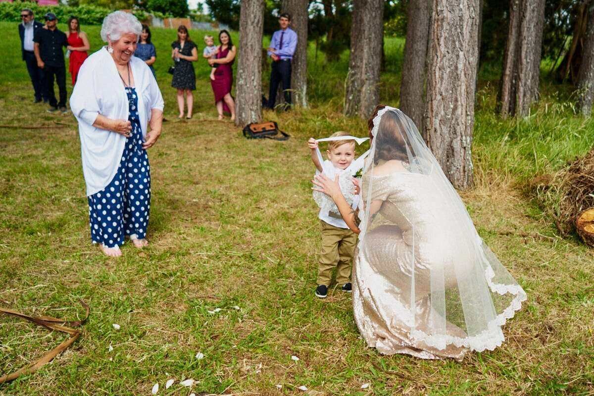 Bride kneeling to hug young boy in suit at outdoor wedding while elderly woman watches on green lawn.